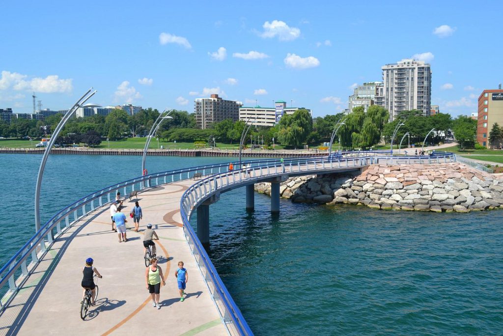 People walk and cycle along a curved waterfront pier beside Lake Ontario, with Burlington’s green shoreline and towers beyond