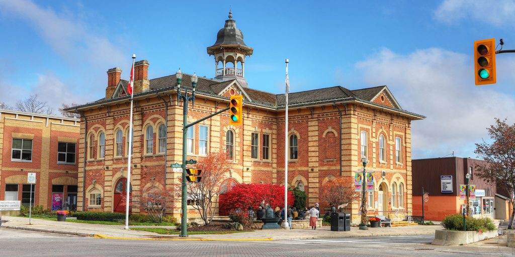Historic brick civic building with a clock tower, ornate windows, mature trees, and traffic lights on an Orangeville street